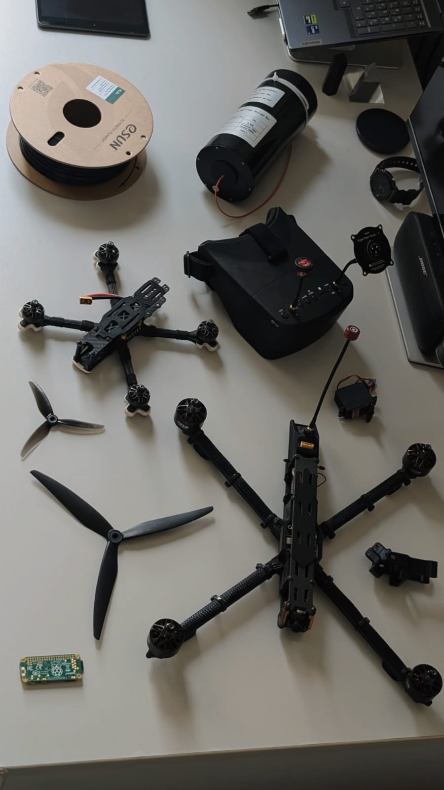 Two quadcopters on a desk: the 10-inch frame with its motors mounted and a smaller quad, with FPV goggles, propellers, a servo and a Raspberry Pi beside them.