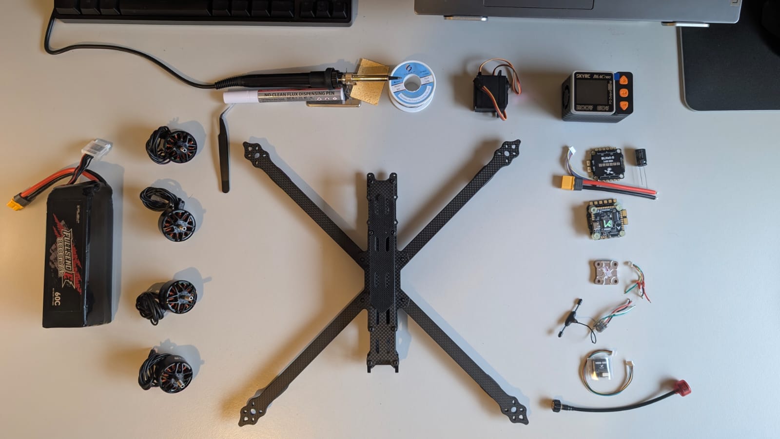 Drone parts laid out on a desk: a carbon fibre X frame, four brushless motors, a LiPo battery, an ESC, a flight controller, a soldering iron and a battery charger.